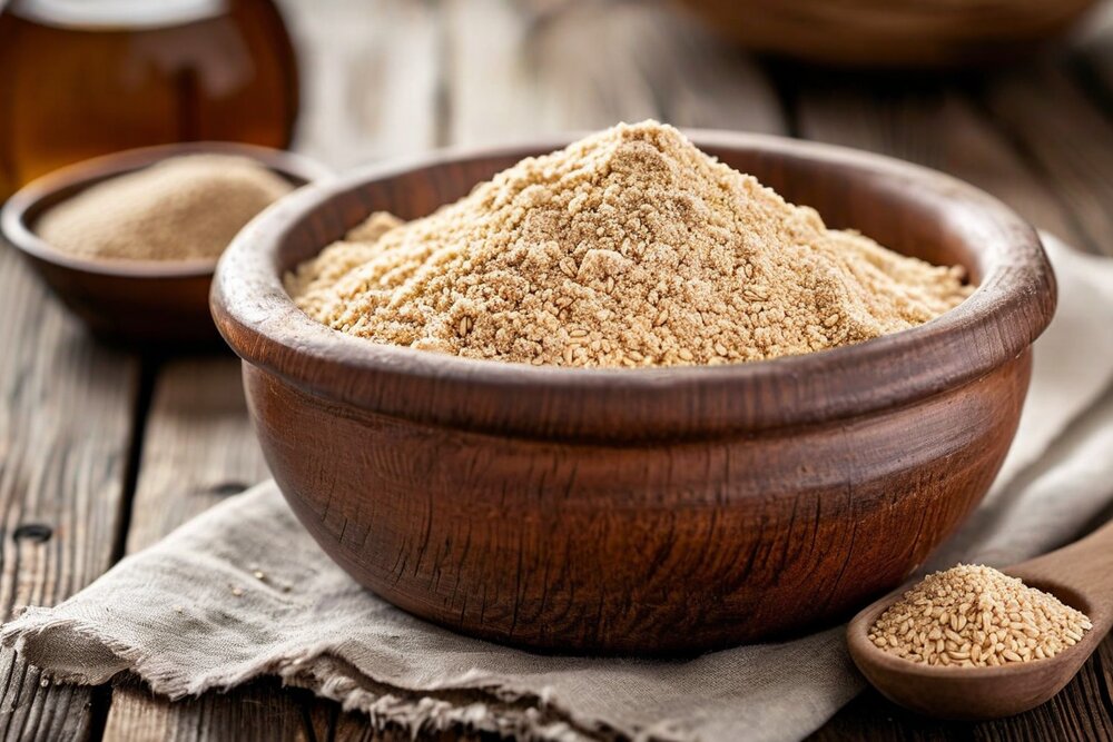 A wooden bowl heaped with pale roasted grain flour, with grains and honey beside it